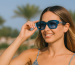 Woman wearing blue OJO sunglasses outdoors, adjusting them while smiling in a sunny, tropical setting.